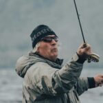 A senior man in a jacket and beanie fly fishing by the sea on a cloudy day.